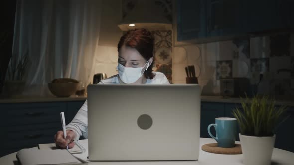 Young Girl In The Kitchen With Laptop, Woman In Medical Mask Makes Notes In Notepad