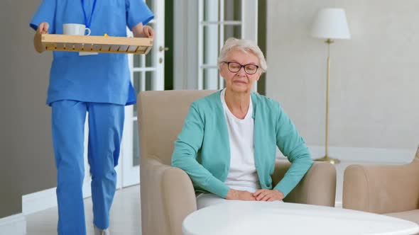 Caretaker carries tray with breakfast and hugs positive single mature grey haired lady