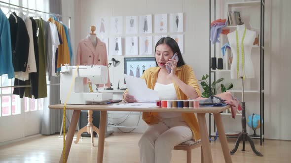 Asian Female Designer With Sewing Machine Talking On Smartphone And Looking At The Paper In Hand