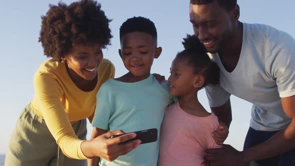 Smiling african american family using smartphone and embracing on sunny beach