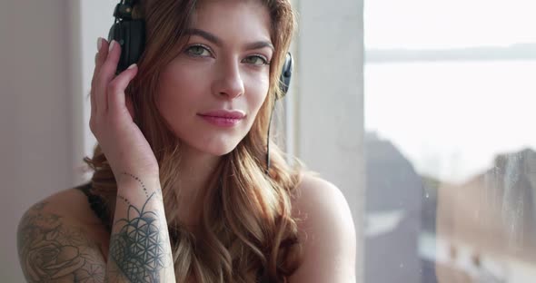 Woman with Headphones on Touching the Earcups and Smiling at Window