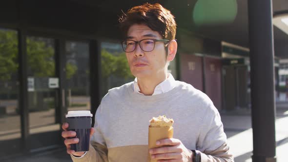 Asian man drinking coffee and having a snack while walking outdoors
