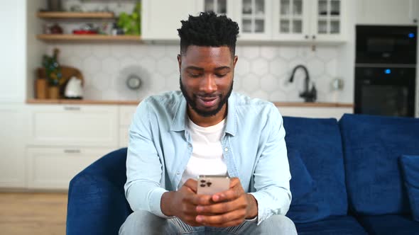 Smiling African American Man Sitting on Sofa Using Mobile Phone for Checking Social Nets