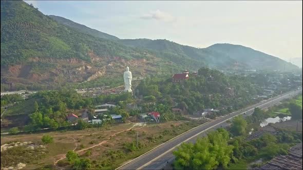 Drone Approaches Across Road To Buddha Statue From Village