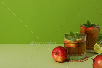 Glasses with apple cider, red apples and piece of lime on green background, space for text