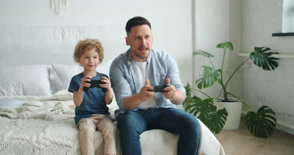 Cute Kid Playing Video Game with Father Holding Joysticks Having Fun in Bed