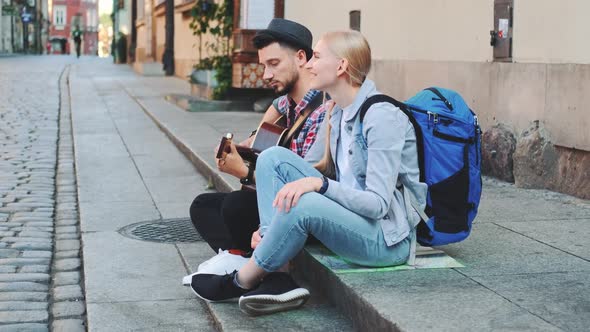 Couple of Tourists Sitting on Sidewalk Playing Guitar and Having Rest