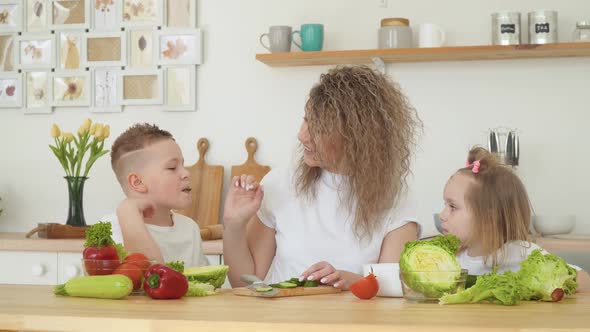 Young Blonde Woman with Curly Hair Gives a Taste of a Slice of Cucumber to Her Son and Daughter