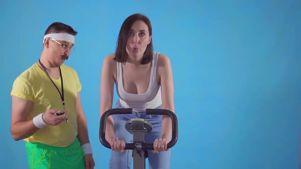 Funny Man Coach From 80's with Mustache and Glasses Examines a Young Woman on Exercise Bike Slow Mo