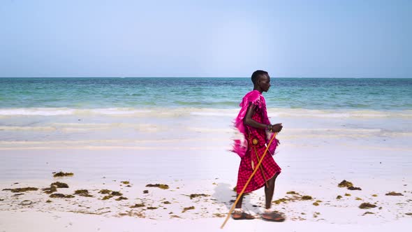 Maasai man in pink clothes walking on sand beach, holding wooden stick.