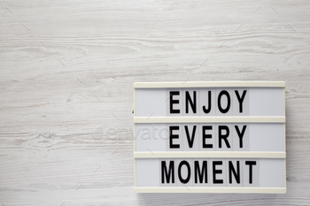'Enjoy every moment' on a lightbox on a white wooden surface, top view.
