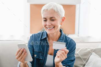 Friendly mature woman using credit debit card and cellphone for e-banking e-commerce at home.