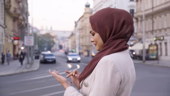 A Young Beautiful Muslim Woman Works on a Smartphone with a Smile in a Street in an Urban Area