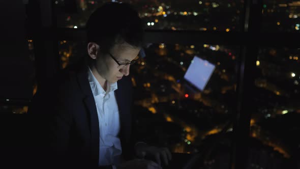 Young Man Freelancer Is Working on His Computer Near the Window in Home at Night.