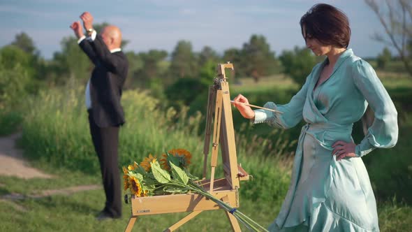 Side View Confident Talented Woman Painting Landscape Outdoors with Blurred Elegant Man Resting