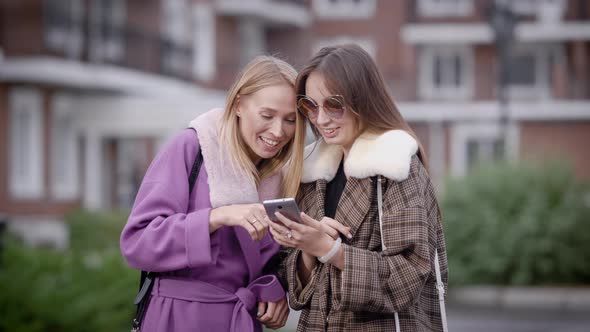 Portrait of Two Girls with a Smartphone Laughing Outdoor in Fall, Girls in Fashionable Clothes