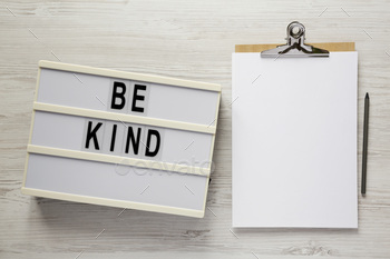 'Be kind' on a lightbox, clipboard with blank sheet of paper on a white wooden surface, top view.