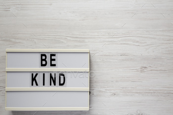 'Be kind' on a lightbox on a white wooden background, top view.