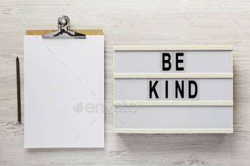 'Be kind' on a lightbox, clipboard with blank sheet of paper on a white wooden background, top view.
