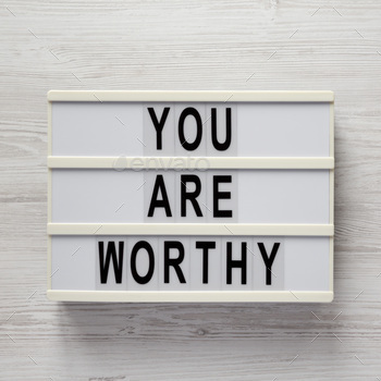 'You are worthy' on a lightbox on a white wooden background, top view. Flat lay, overhead.