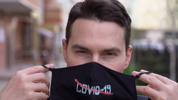 Young brunette man wear black jacket, put on face mask with cross out red line inscription Covid-19