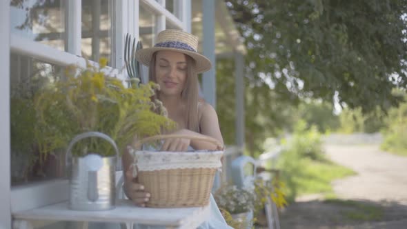 Cute Young Woman in a Straw Hat and White Dress Taking Apple From a Small Basket Sitting in Front