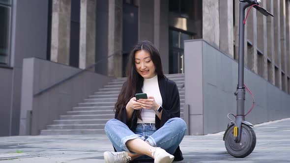 Asian Girl Sitting on the Concrete Ground Near Own e-Scooter on the Beautiful Urban Building
