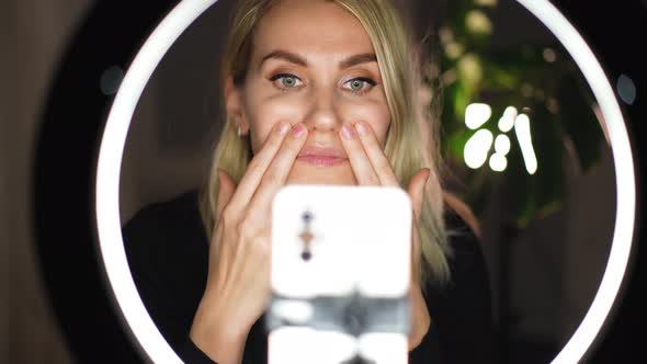 Adorable woman streaming beauty vlog on mobile phone with ring lamp from home