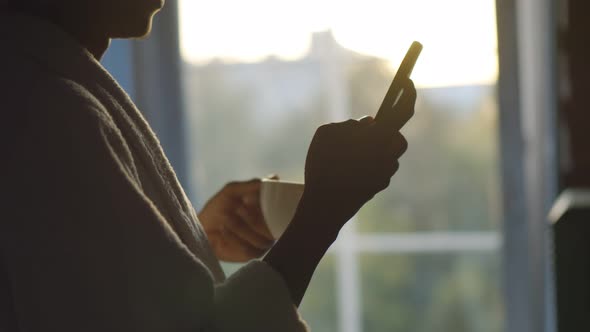 Close Up of Young Afro Man in Bathrobe Drinking Coffee and Texting Sms on Smartphone Near Window