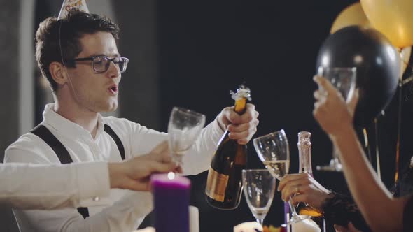 Millennial Friends at Party Sitting at Table, Opening Bottle of Champagne and Pouring Sparkling Wine