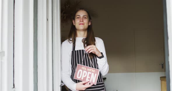 Smiling caucasian waitress standing in door, holding open sign, looking at camera