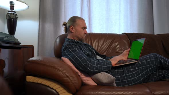 Side View Focused Man in Pajamas with Chromakey Laptop Typing on Keyboard Lying on Couch