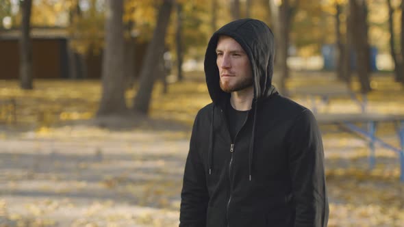 Young Positive Caucasian Man Wearing Hoodie Standing in Autumn Urban Park and Smiling to Camera