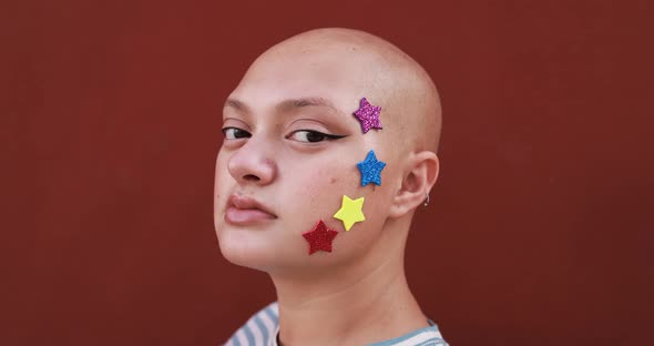 Bald girl with colorful stars on her face posing in front of camera with red background