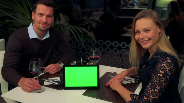 A Man and a Woman Sit at a Table in a Restaurant and Smile Into Camera, a Tablet with a Green Screen