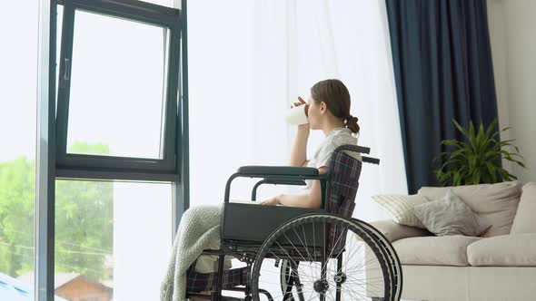 Young Woman Sitting on the Wheelchair Reading a Book and Drinking Coffee at Home