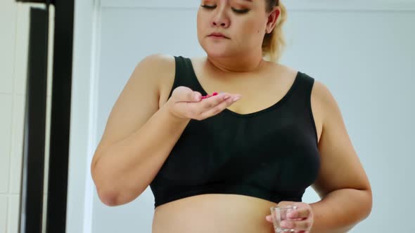 Overweight woman holding diet pills and looking her belly fat into the mirror