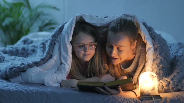 Mom and Daughter Reading Book in Bed Under Blanket