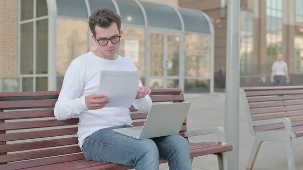 Young Man Reading Documents and Working on Laptop While Sitting on Bench Outdoor