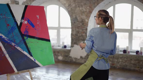 Woman Uses Wide Brush and Color Paints Splashing Gesture on Canvas in Artstudio