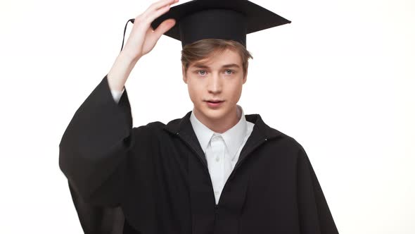 Young Caucasian Graduate in Black Robe Rearranging His Square Academic Cap on White Background