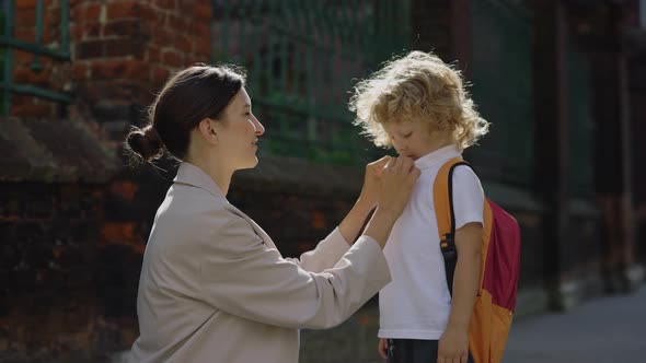 Young Mother Preparing and Caring Her Little Curly Son to School Outside