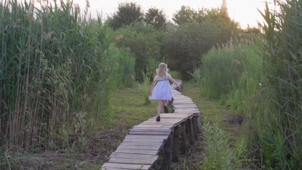 Cheerful Active Little Girl in White Dress Runs Along Wooden Bridge Outdoors Among Green High Reeds
