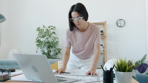 Young Female Architect Working at Building Plan Drawing on Blueprint and Using Laptop at Home