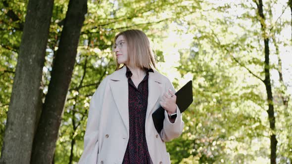 Young Girl Walks in the Park on a Sunny Day Wearing Coat and Drinking From a Cup