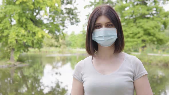 A Young Caucasian Woman in a Face Mask Shows a Thumb Up to the Camera and Nods in a Park