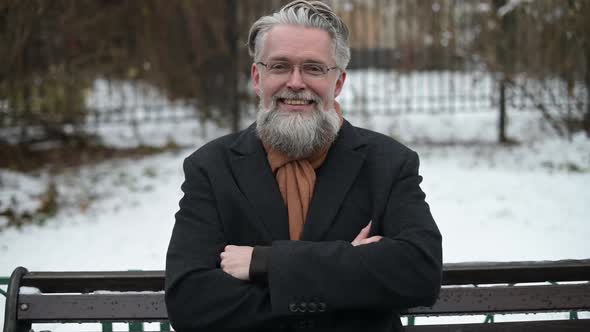 A respectable gray-haired man in glasses sits on a park bench