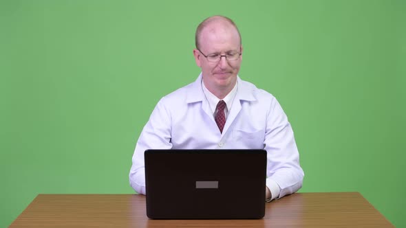 Stressed Mature Bald Man Doctor Arguing To the Laptop Against Wooden Table