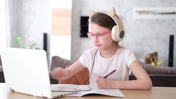 A Smiling Little Caucasian Girl with Headphones Looks at Her Laptop Sitting at the Table Listening