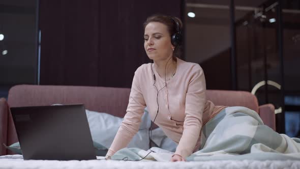 Wide Shot Caucasian Woman in Headphones Surfing Internet on Laptop Sitting on Bed at Home